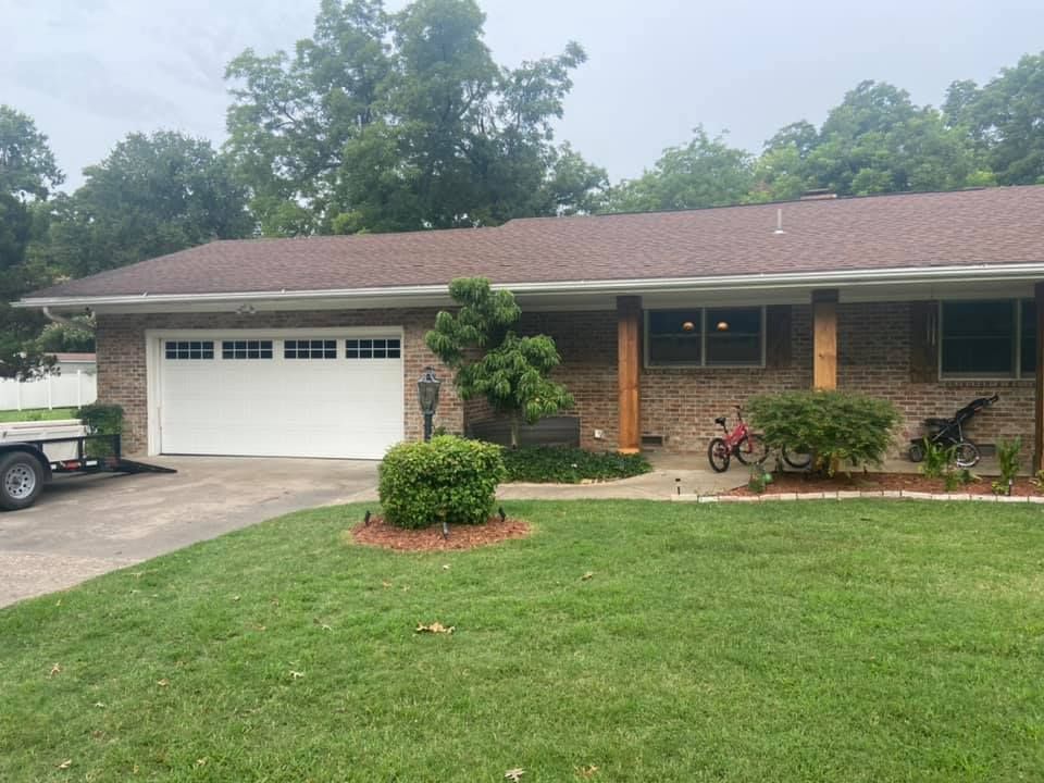 Ranch-style house with white garage door, brown brick exterior, and green lawn. A small trailer is parked to the left.
