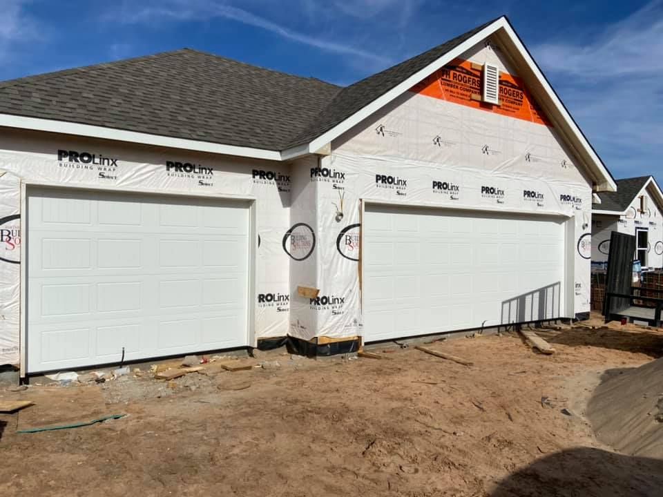 Two-car garage under construction with white doors, wrapped in white material, against a blue sky.