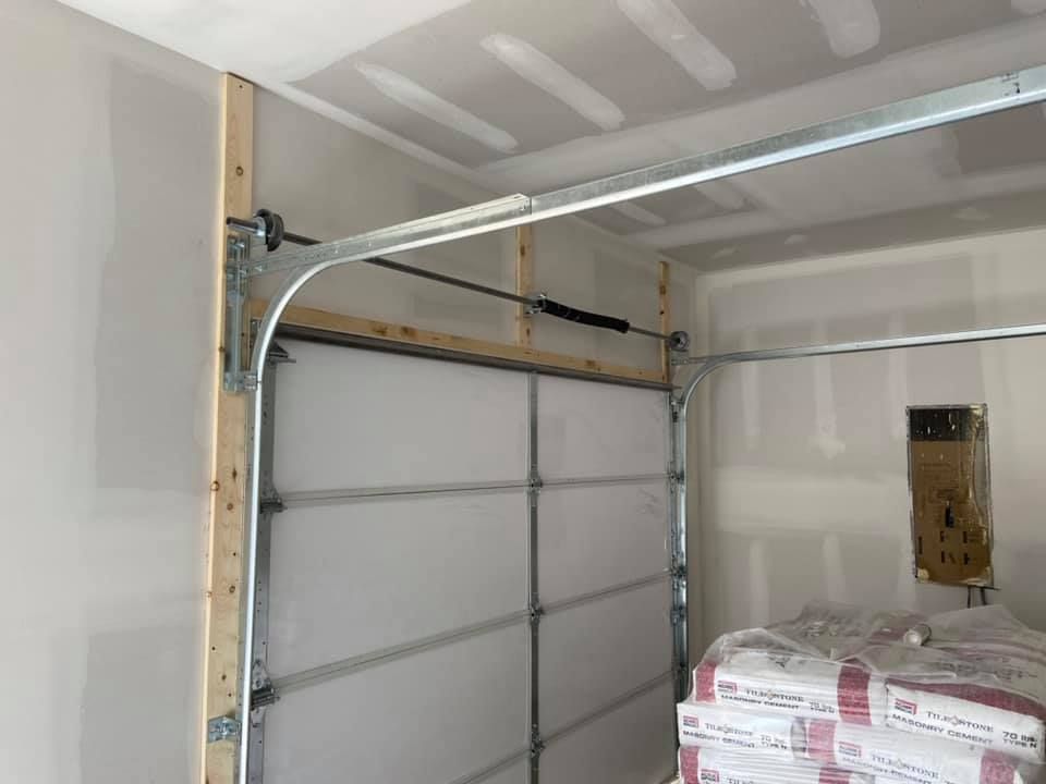 Garage door partially open, inside a room with drywall. A mechanical track and spring are visible.