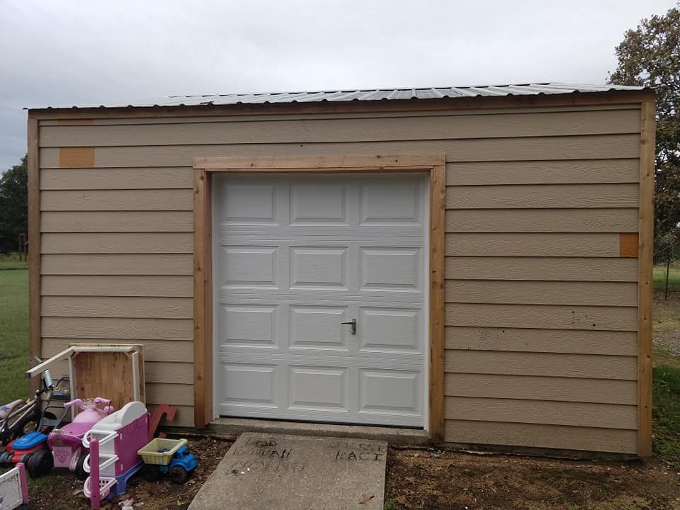 Tan shed with white garage door and wood trim, small items sit at the base, under a cloudy sky.
