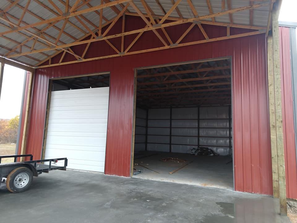 Red metal barn with open bays; a white sliding door on the left, a trailer in foreground.