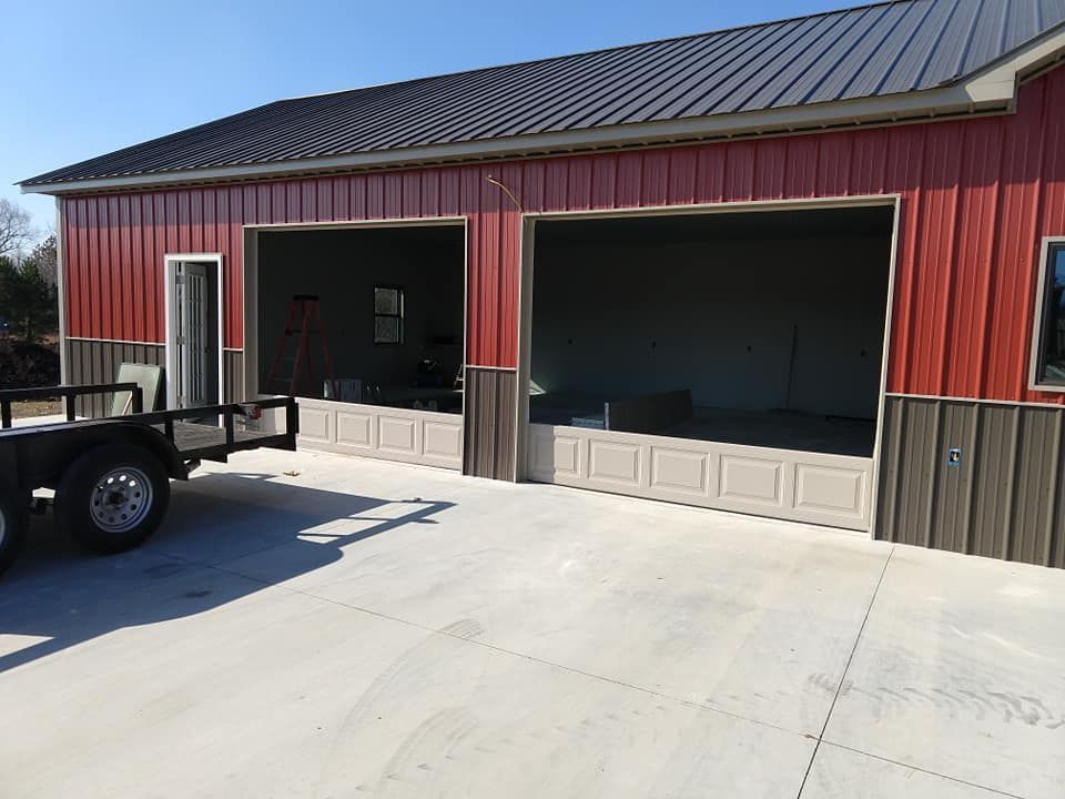 Red and brown metal-clad garage with open bays. A trailer is parked on the concrete driveway.