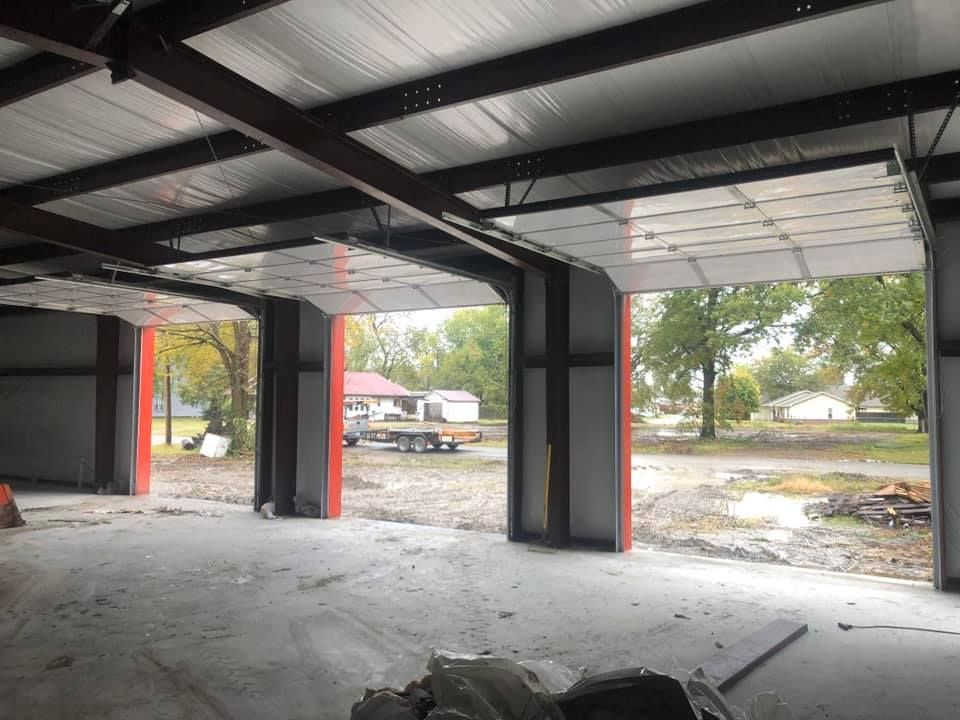 Inside a steel-framed building with three open garage door openings, revealing an exterior view.