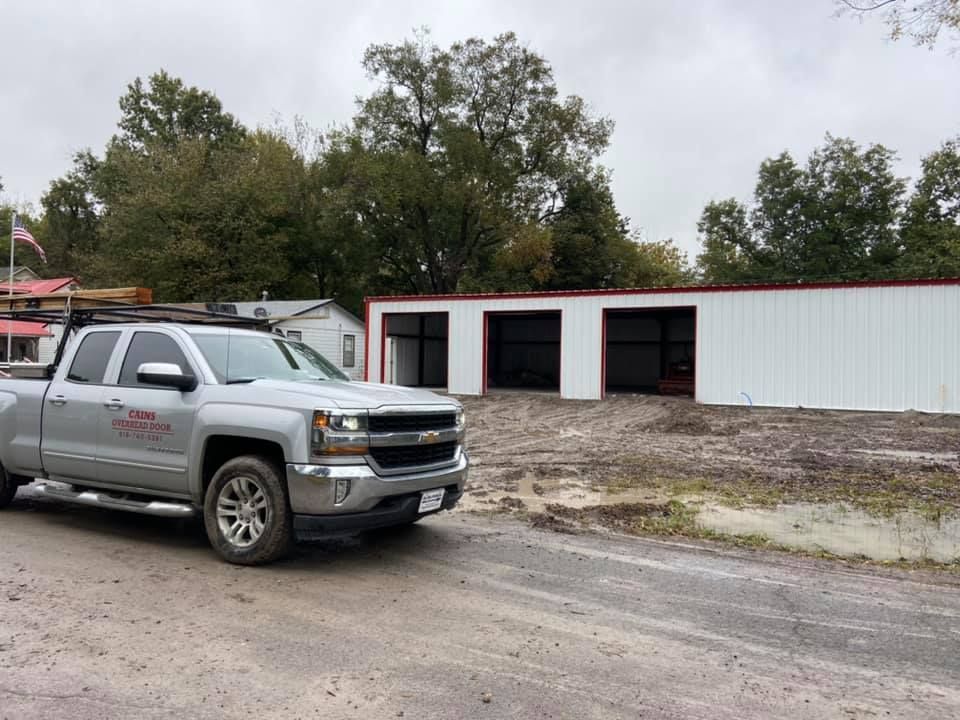 Silver truck parked near a newly built white and red metal building with three open bays.
