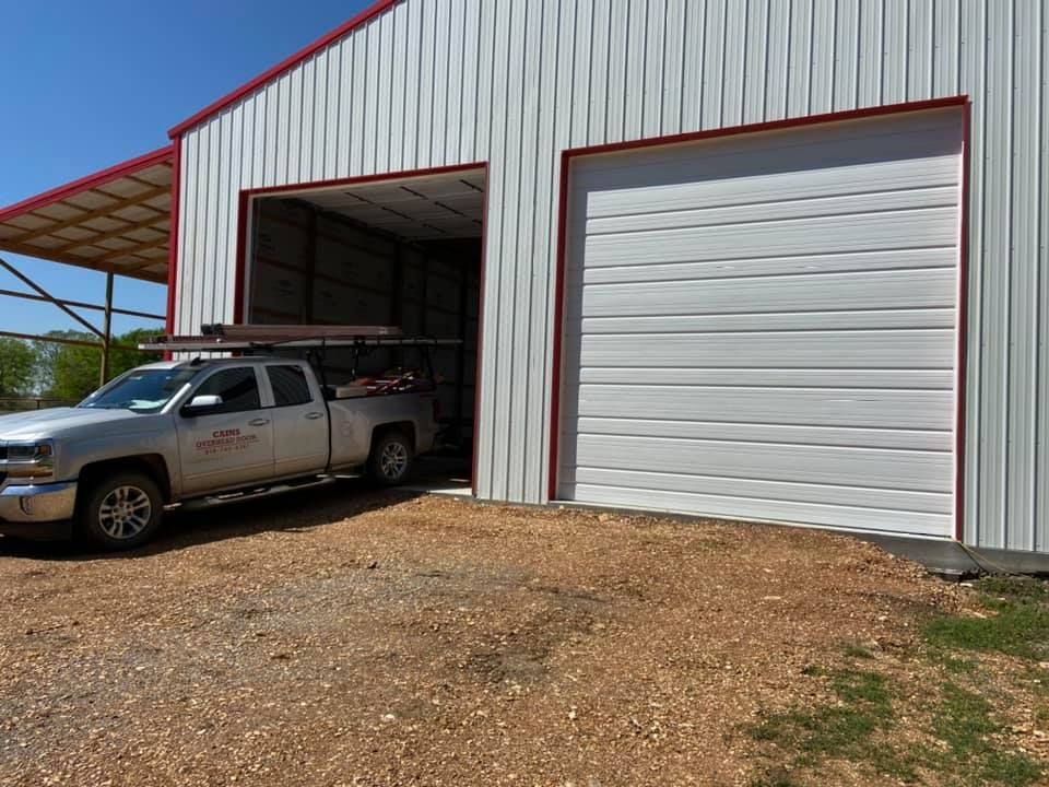 Silver pickup truck parked in front of a white building with two large garage doors. Red trim and a blue sky.