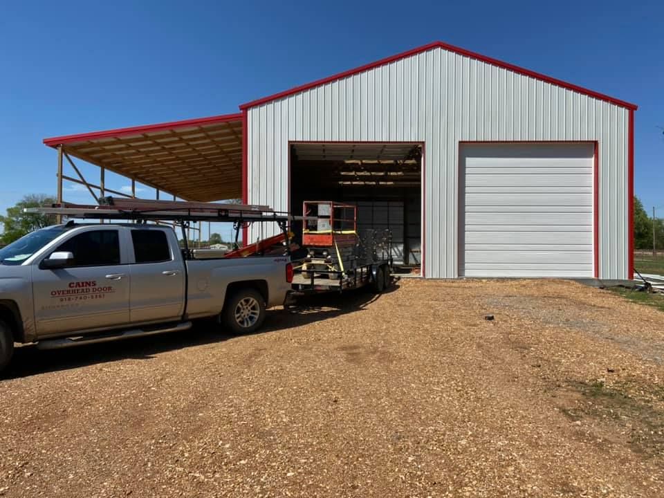Silver truck with trailer in front of a white metal building with red trim.
