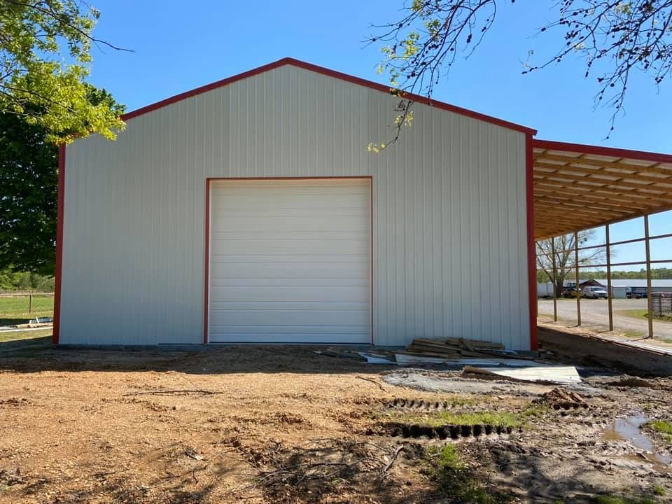White metal barn with red trim, a large garage door, and a carport in a dirt lot.