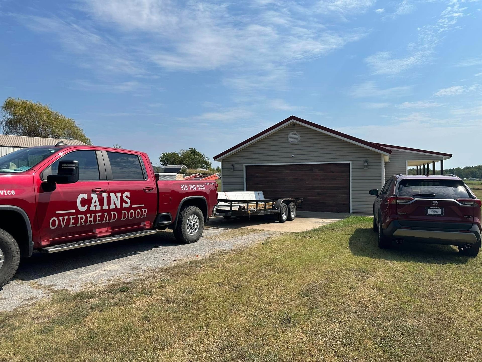 Red pickup truck with a trailer parked in front of a garage. A car is parked to the right. Sunny day.