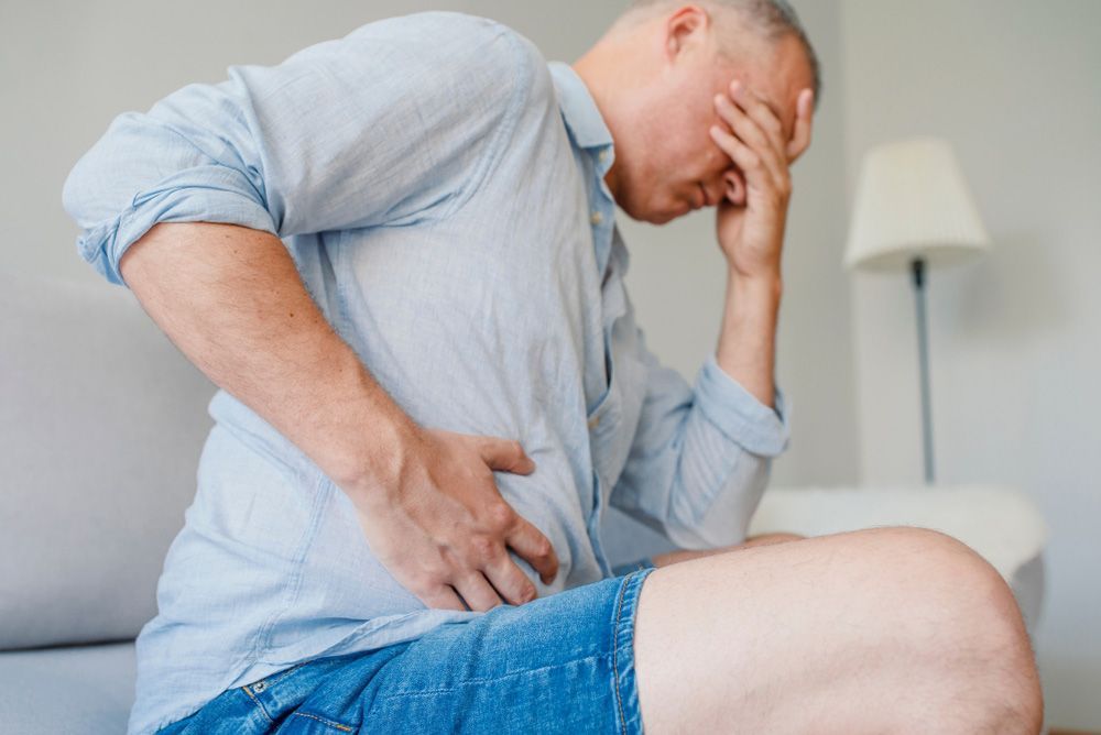 A Man is Sitting on a Couch Holding His Stomach in Pain — Brisbane Upper GI Surgery in Chermside, QLD