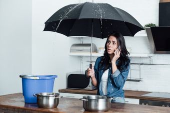 Woman in kitchen holding umbrella indoors, talking on phone as water leaks from ceiling into pots and bucket.