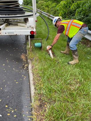 A man in a yellow vest is kneeling down in the grass next to a road.