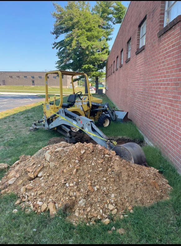 A yellow tractor is digging a hole in the ground in front of a brick building.