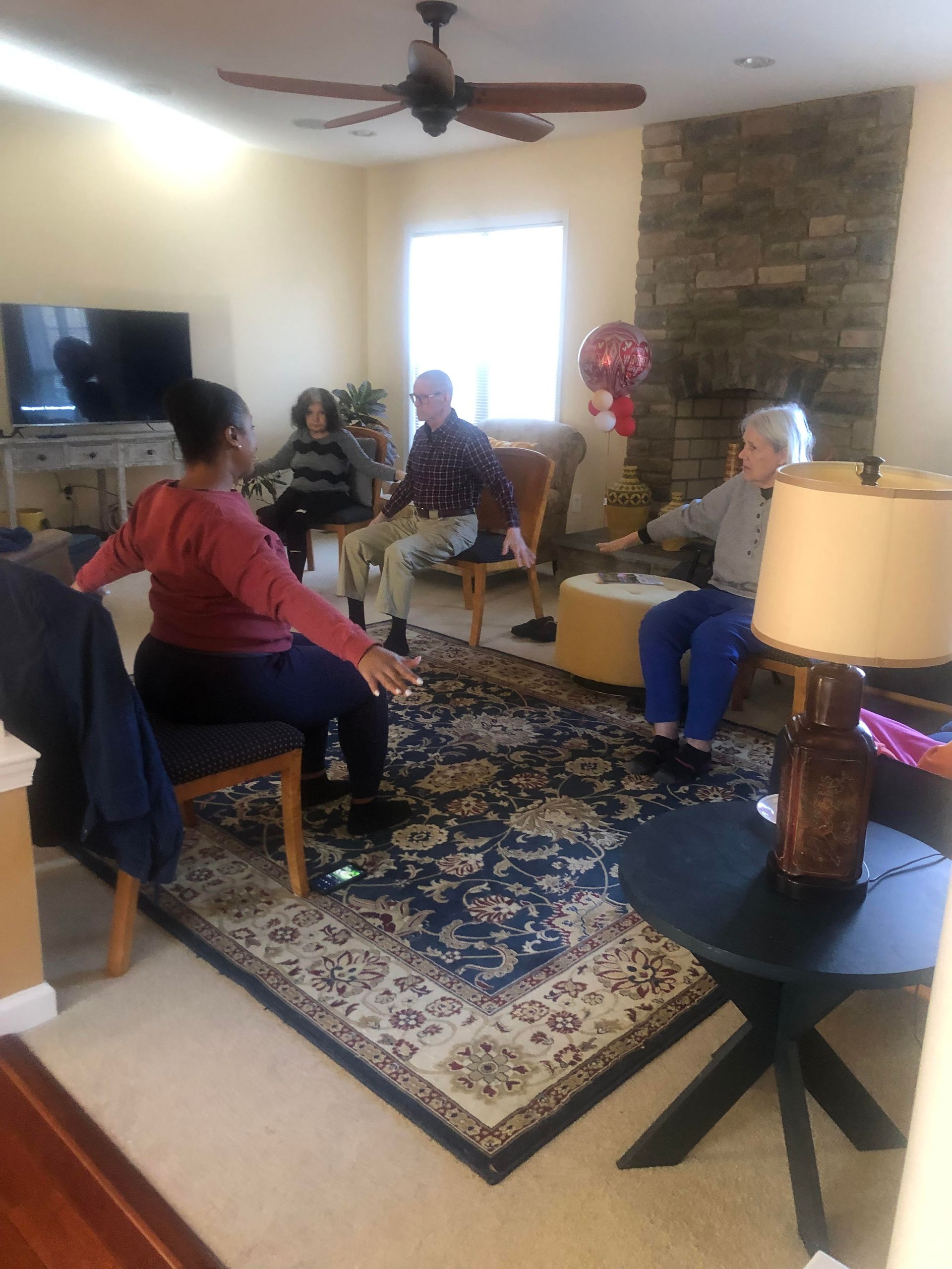 Four people sit on chairs in a living room, performing seated arm-stretching exercises together.