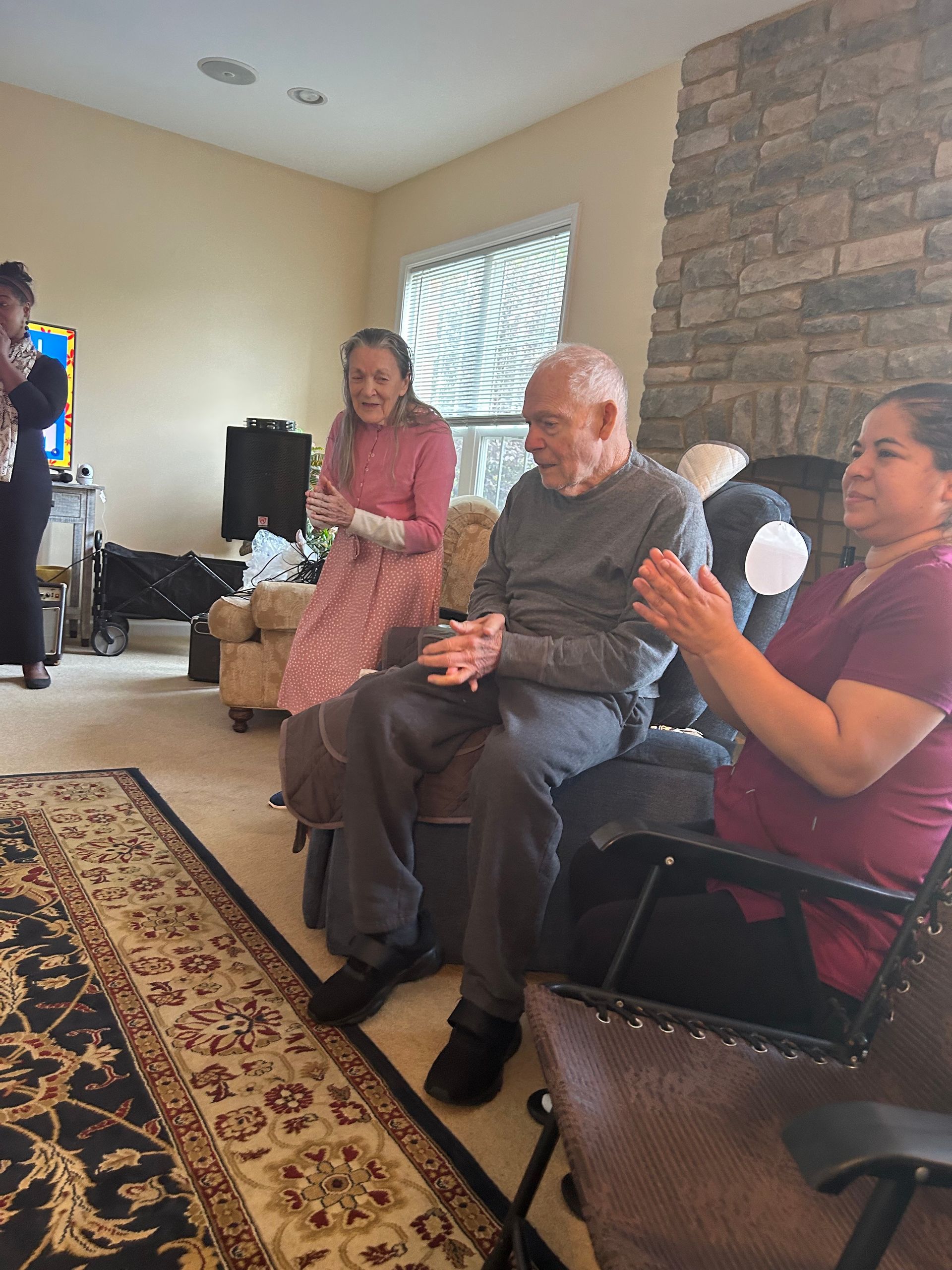 A group of people sitting and standing in a living room, smiling and clapping near a stone fireplace.