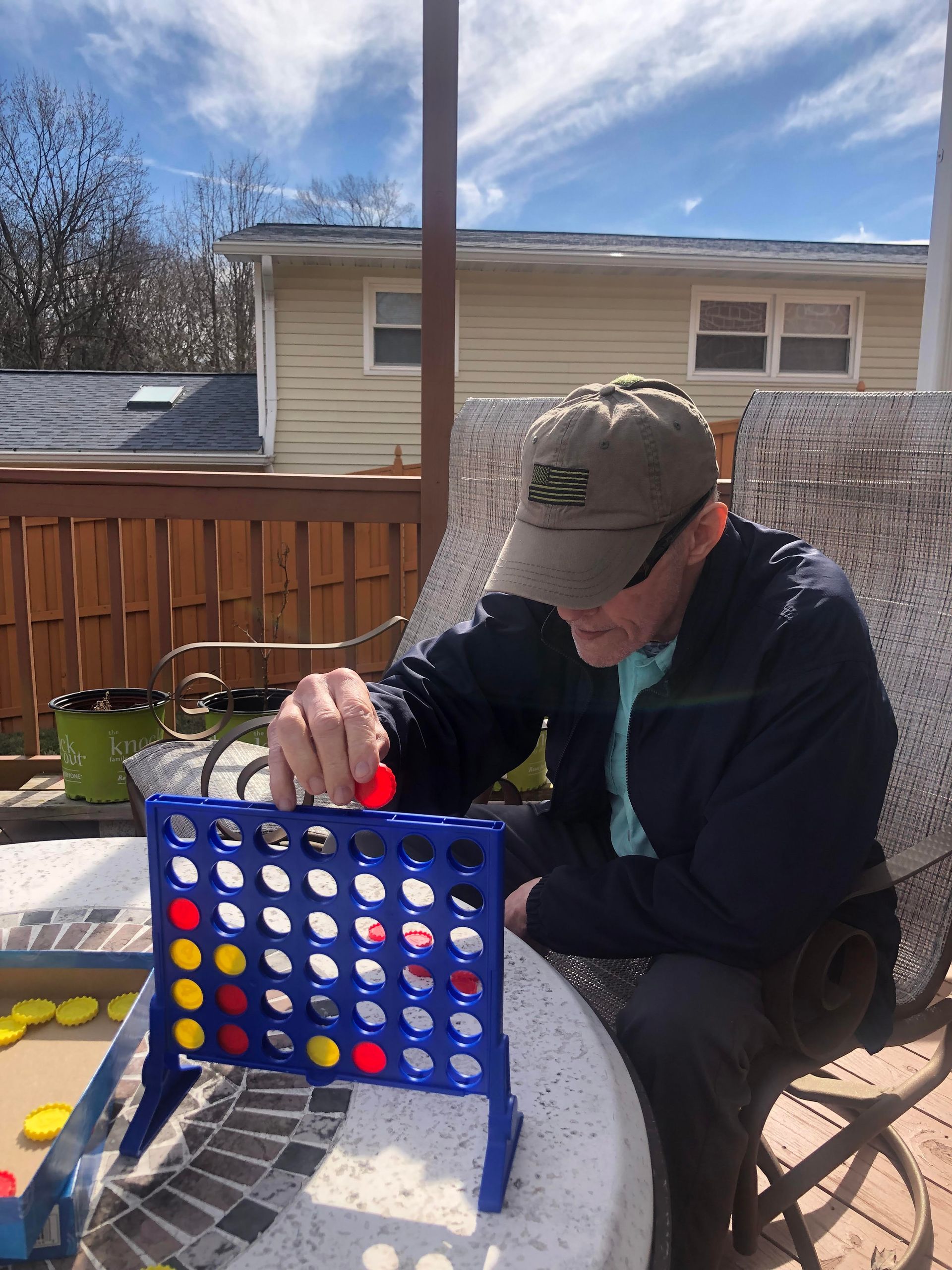 An adult in a baseball cap plays Connect Four on a patio table with yellow and red game pieces.
