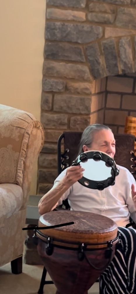 An individual sits indoors playing a djembe drum while holding a tambourine in front of a stone fireplace.