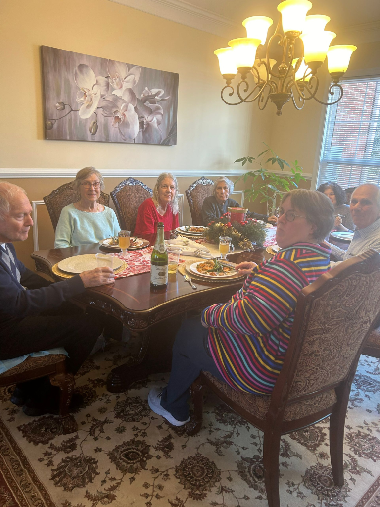 Group of people seated around a table, dining. Food and drinks are present. A chandelier hangs overhead.