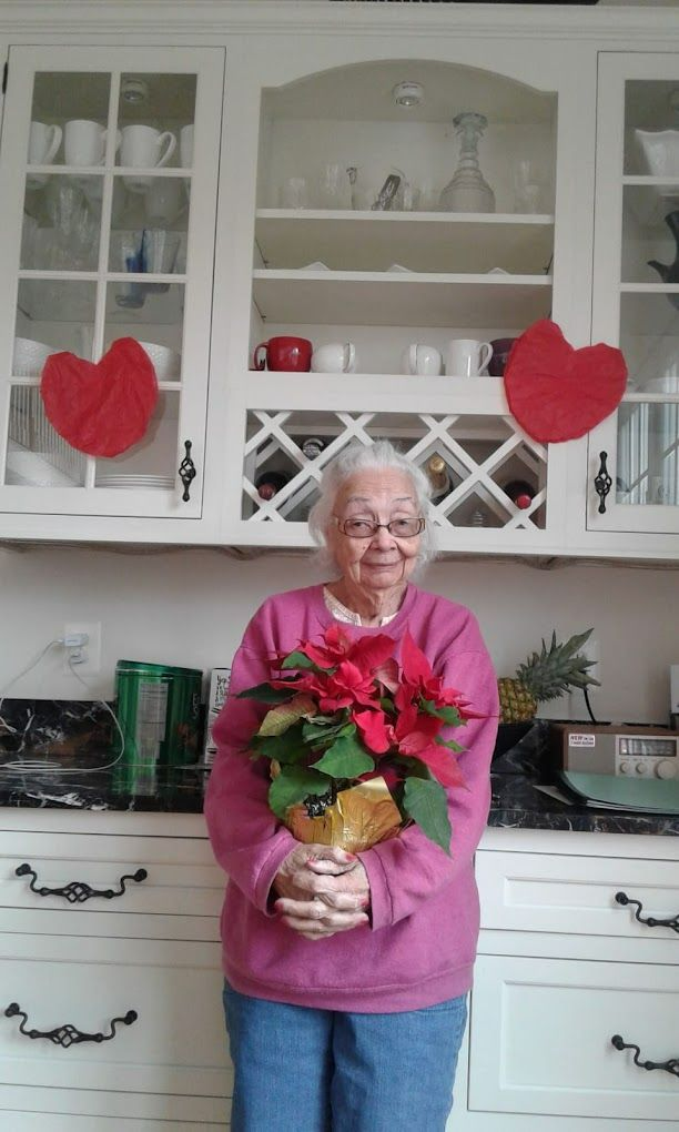 Woman holding poinsettia, smiling, in front of white cabinets with red heart decorations.