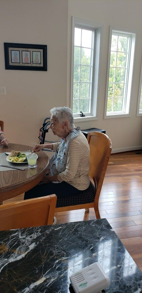 An elderly person sitting at a table near a window. There are plates of food.