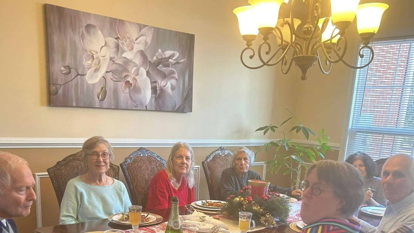 People seated at a dining table, possibly a holiday gathering. A chandelier and floral artwork are visible.