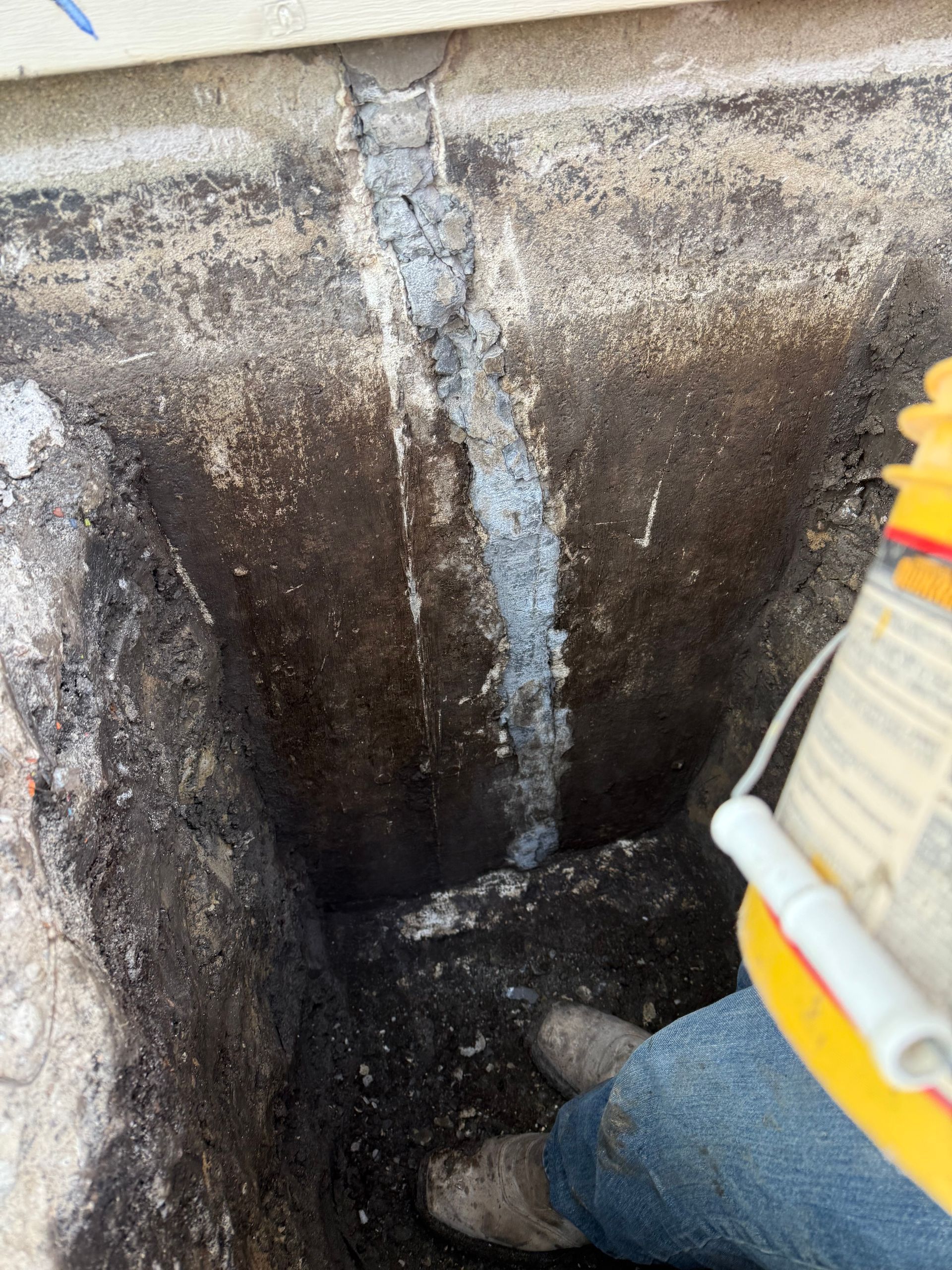 A person in muddy boots kneels in a trench, holding a yellow container. A vertical, cement-filled crack runs up a wall.