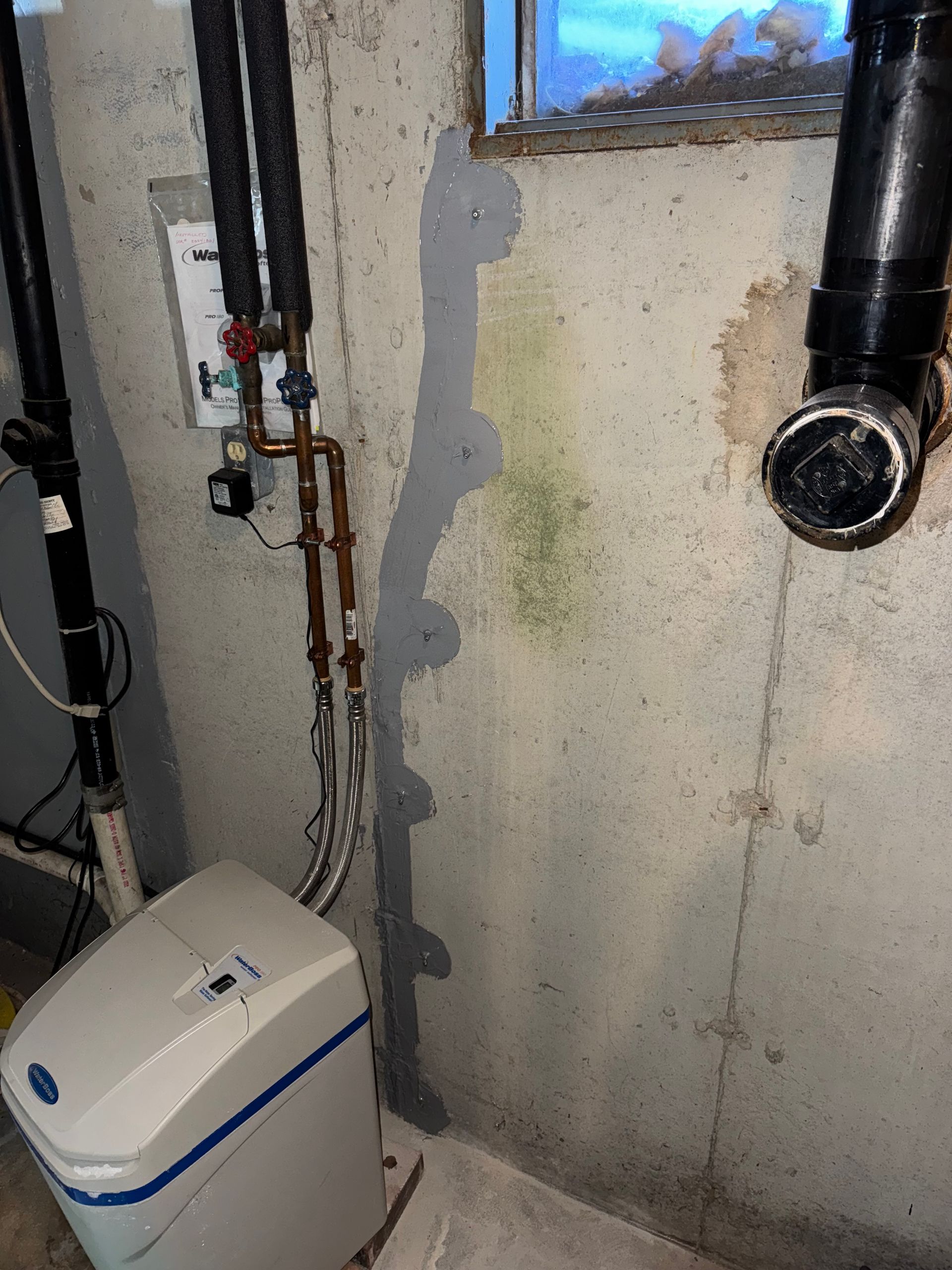 Basement wall with a water leak, visible by the gray sealant and greenish mold. A water softener and pipes are also present.