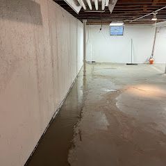 Flooded basement with water along the wall and on the floor. White walls, ceiling, and support beams are visible.