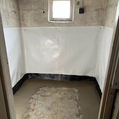 A small, unfinished basement with white waterproof sheeting on the walls, a concrete floor, and a small window.