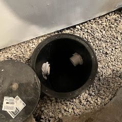 Open black pit with a partially removed lid, surrounded by gravel, indoors. Two white pipes are visible inside.