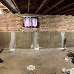 Basement wall with patched concrete and a small window with curtains. Overhead, wooden beams. Brick wall at the top.