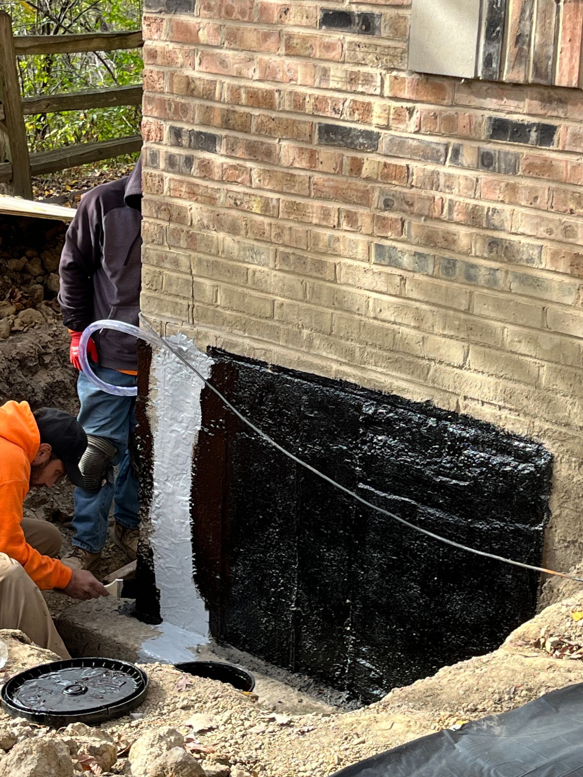 Two people applying black sealant to a brick foundation wall during construction. One sprays the wall with sealant, while the other smoothes it.