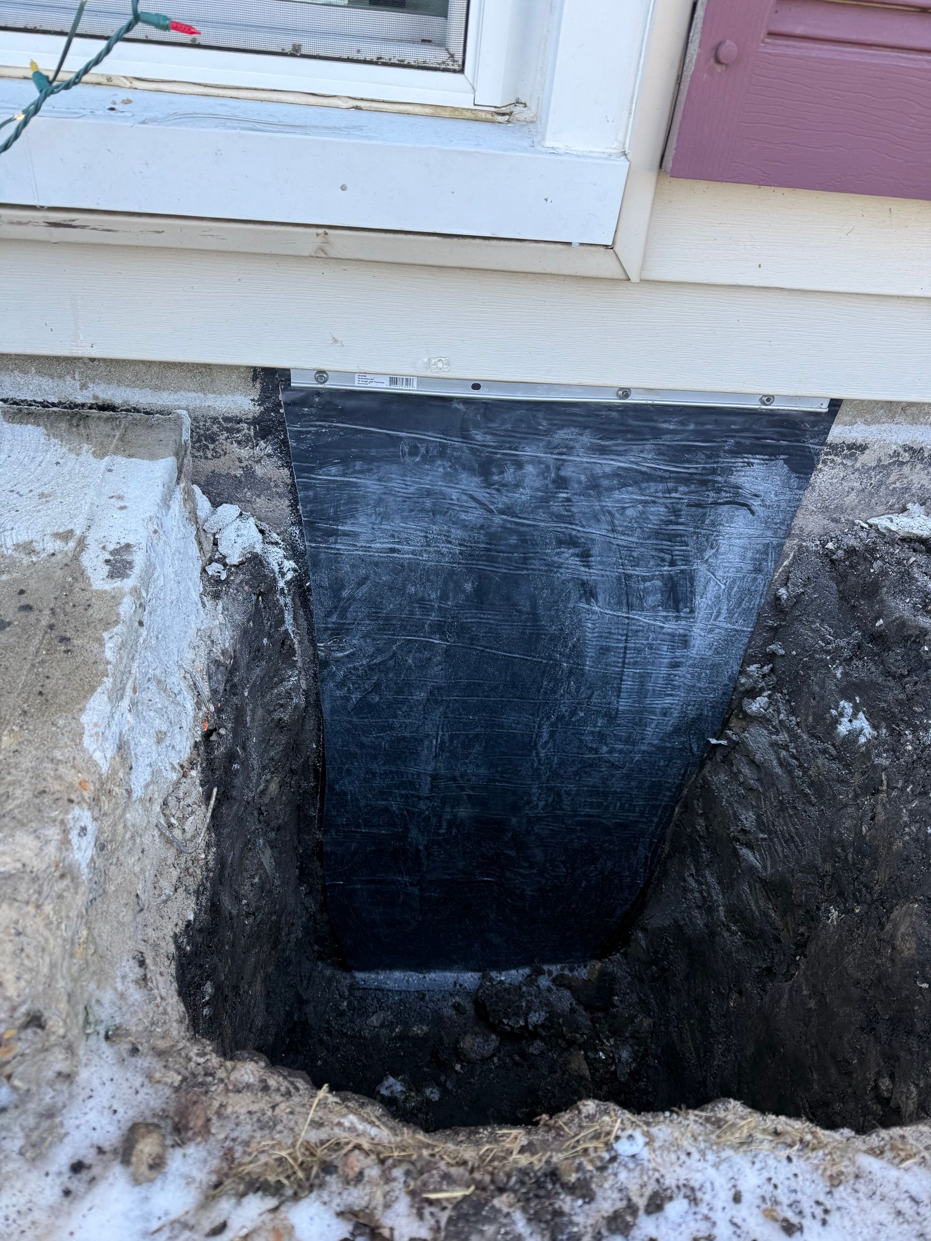 A rectangular hole dug next to a house foundation, revealing a black surface, likely waterproofed. Snow is visible nearby.