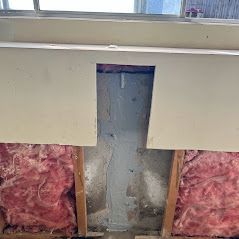 Wall with a rectangular hole revealing insulation and wooden studs. Pink insulation is visible on either side of the hole.