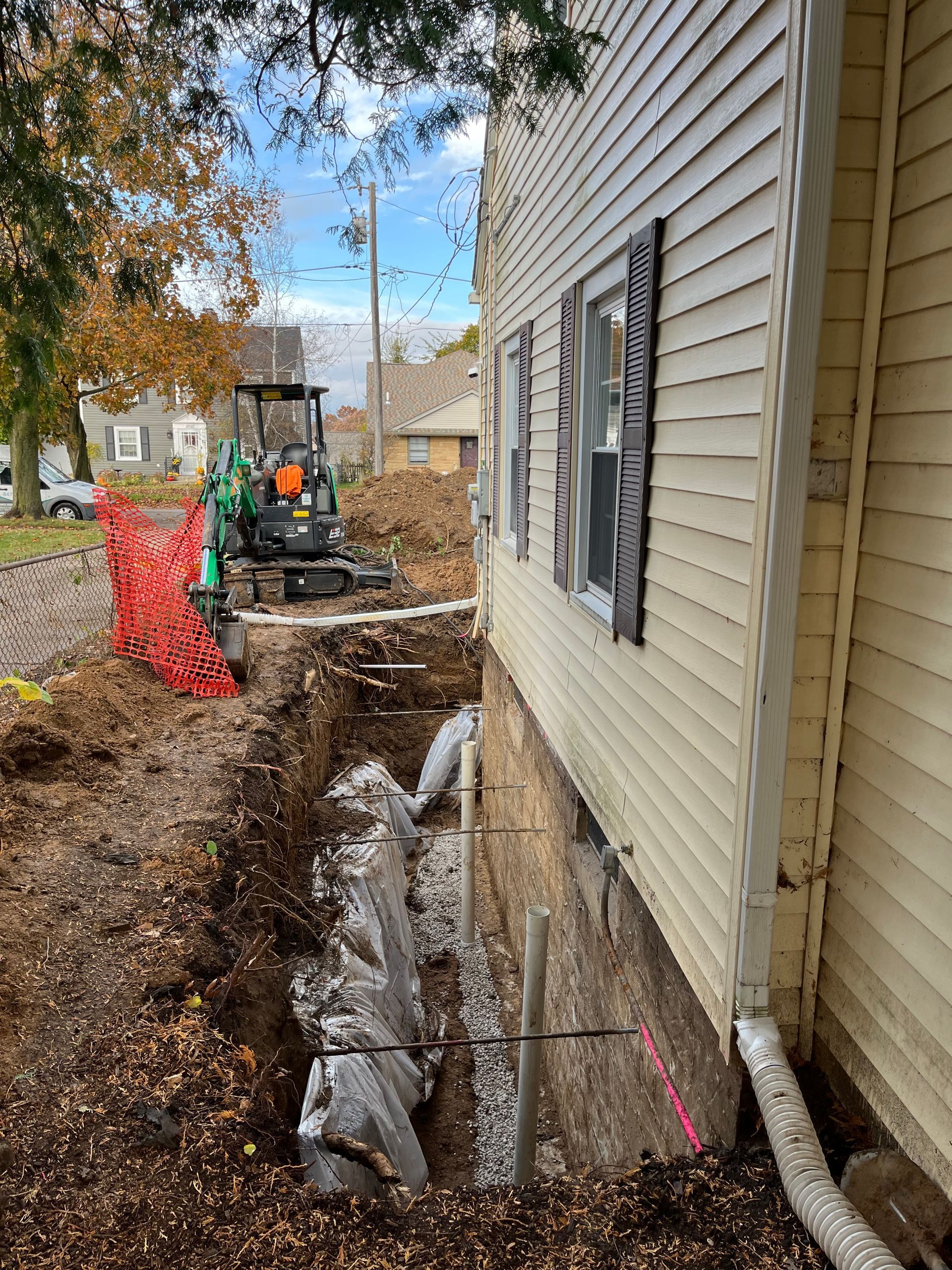 A trench is dug alongside a house with yellow siding. A small excavator and orange safety fencing are also present.