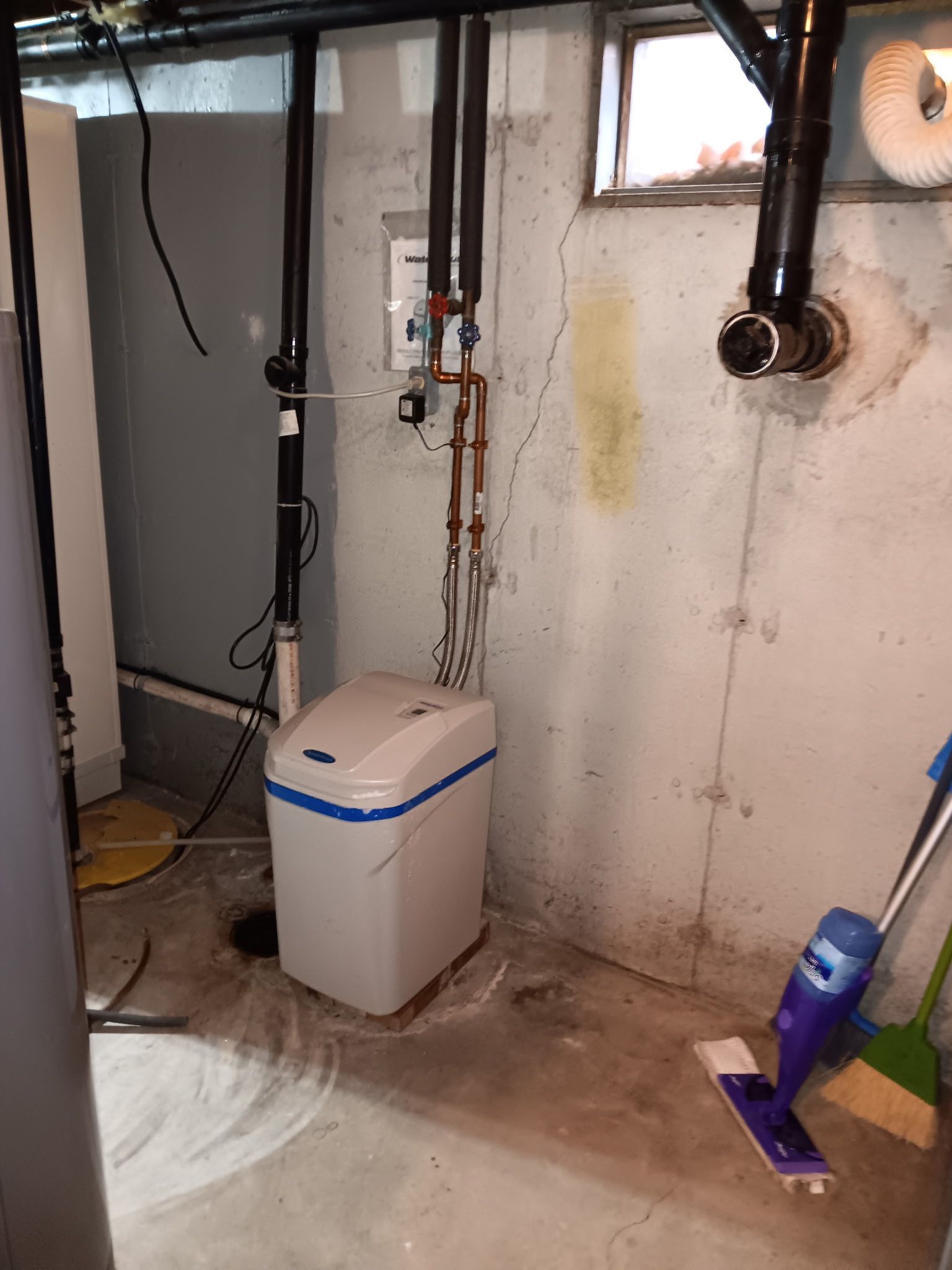 A water softener in a basement. White tank with blue trim, copper pipes, and a gray wall. Mop and broom on the floor.