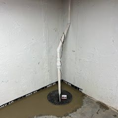 A sump pump system in a basement corner. A white pipe runs up the wall from a circular base.
