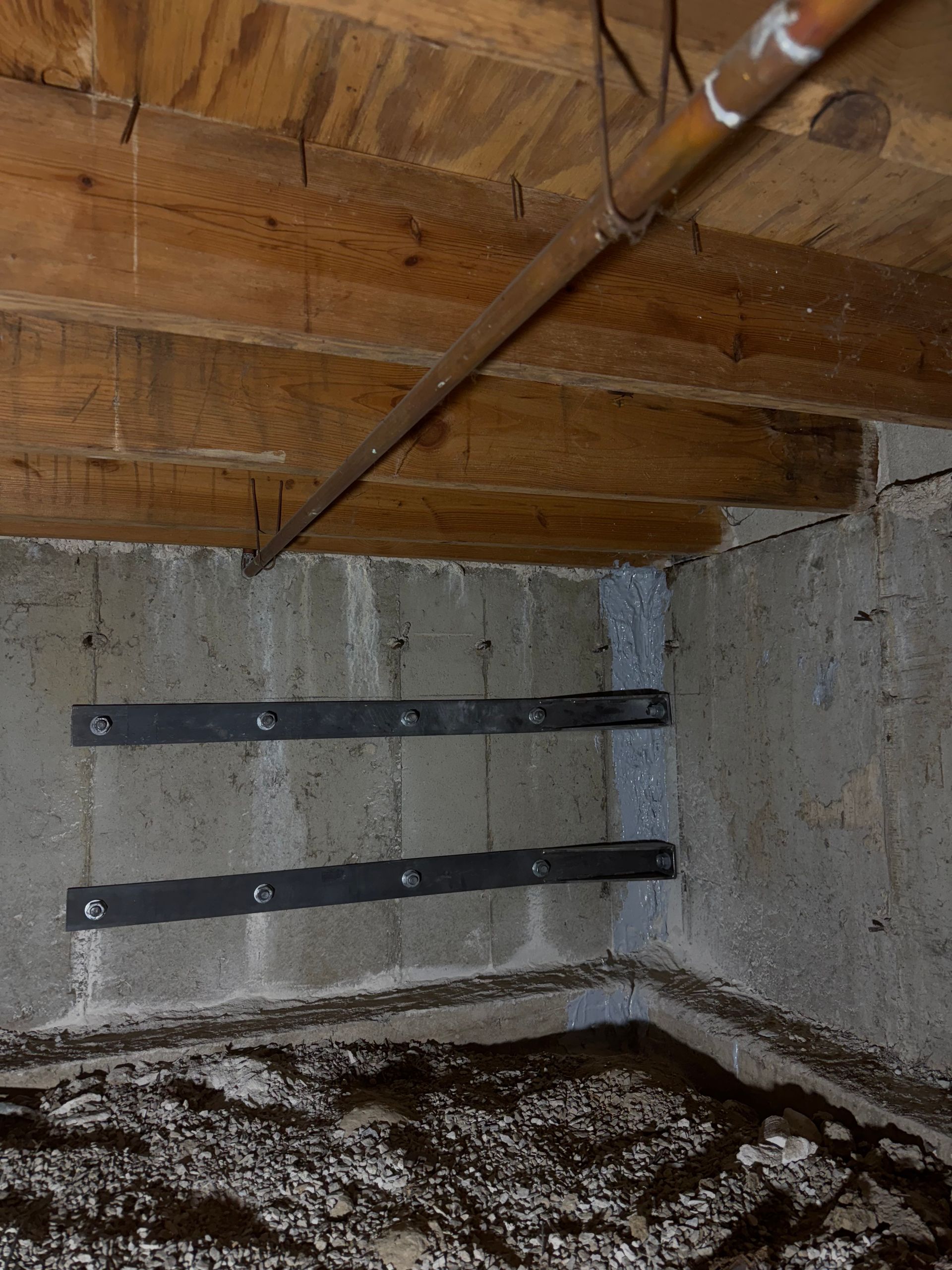 Two black metal reinforcement straps bolted to a gray concrete basement wall, under wooden beams.
