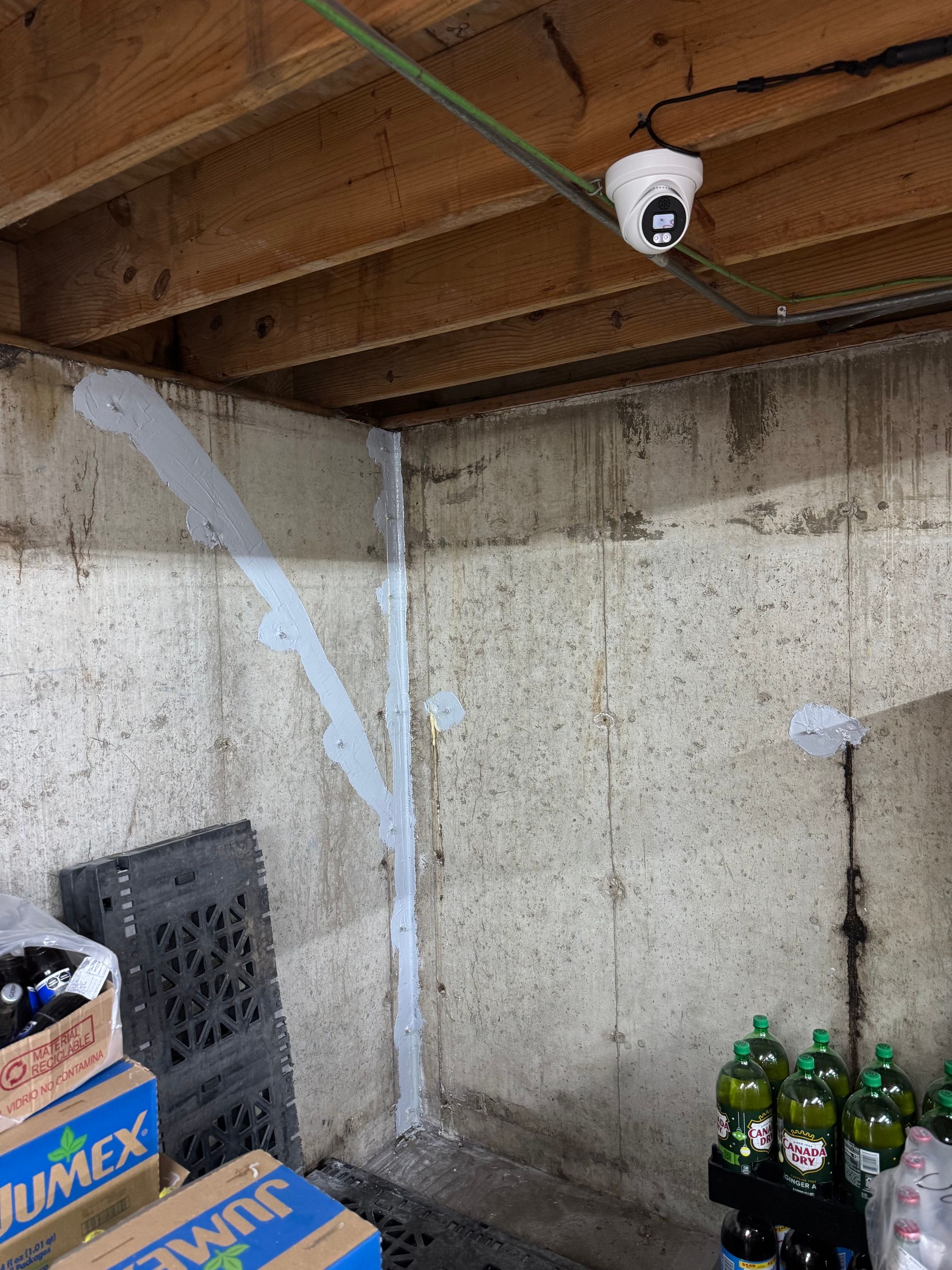 Security camera mounted on a wooden ceiling in a basement corner. Concrete walls show a light-colored sealant, with crates and bottles below.