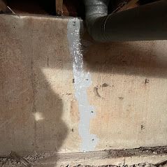 Gray sealant applied to a concrete basement wall, near a pipe.  Several fasteners are embedded in the sealant.
