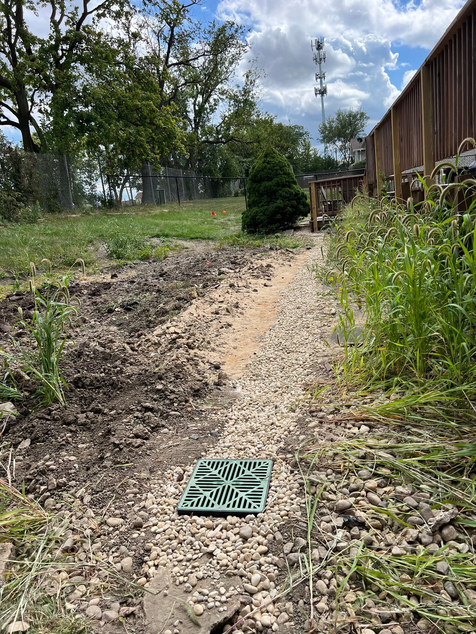 A gravel-lined trench leads toward a green drain cover. Vegetation grows along the sides, with a fence and trees in the background.
