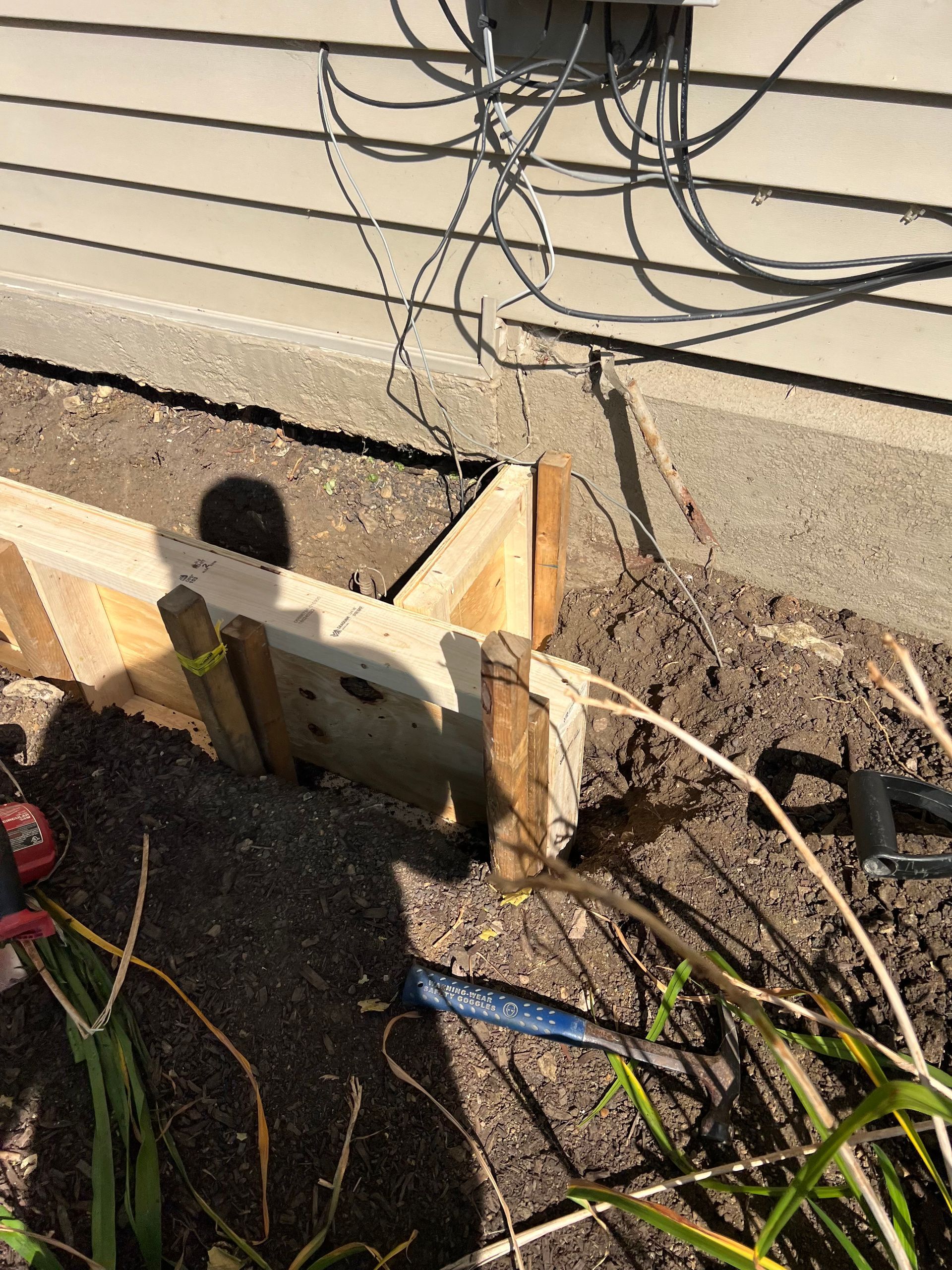Wooden formwork next to a building foundation, set in soil, with wiring visible above.