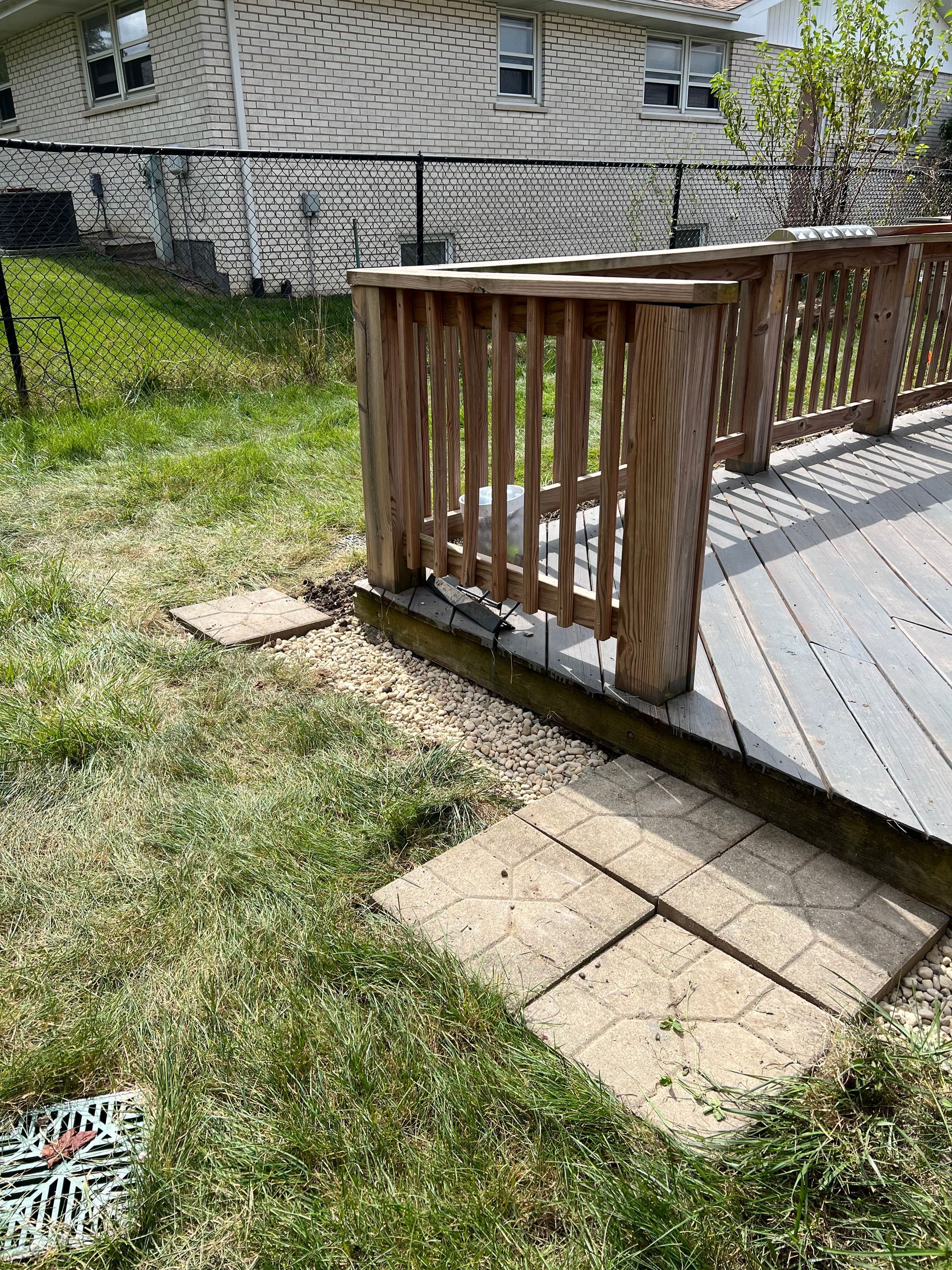 A wooden deck with a railing is next to a grassy lawn with a pathway of concrete squares leading away. A house is in the background.