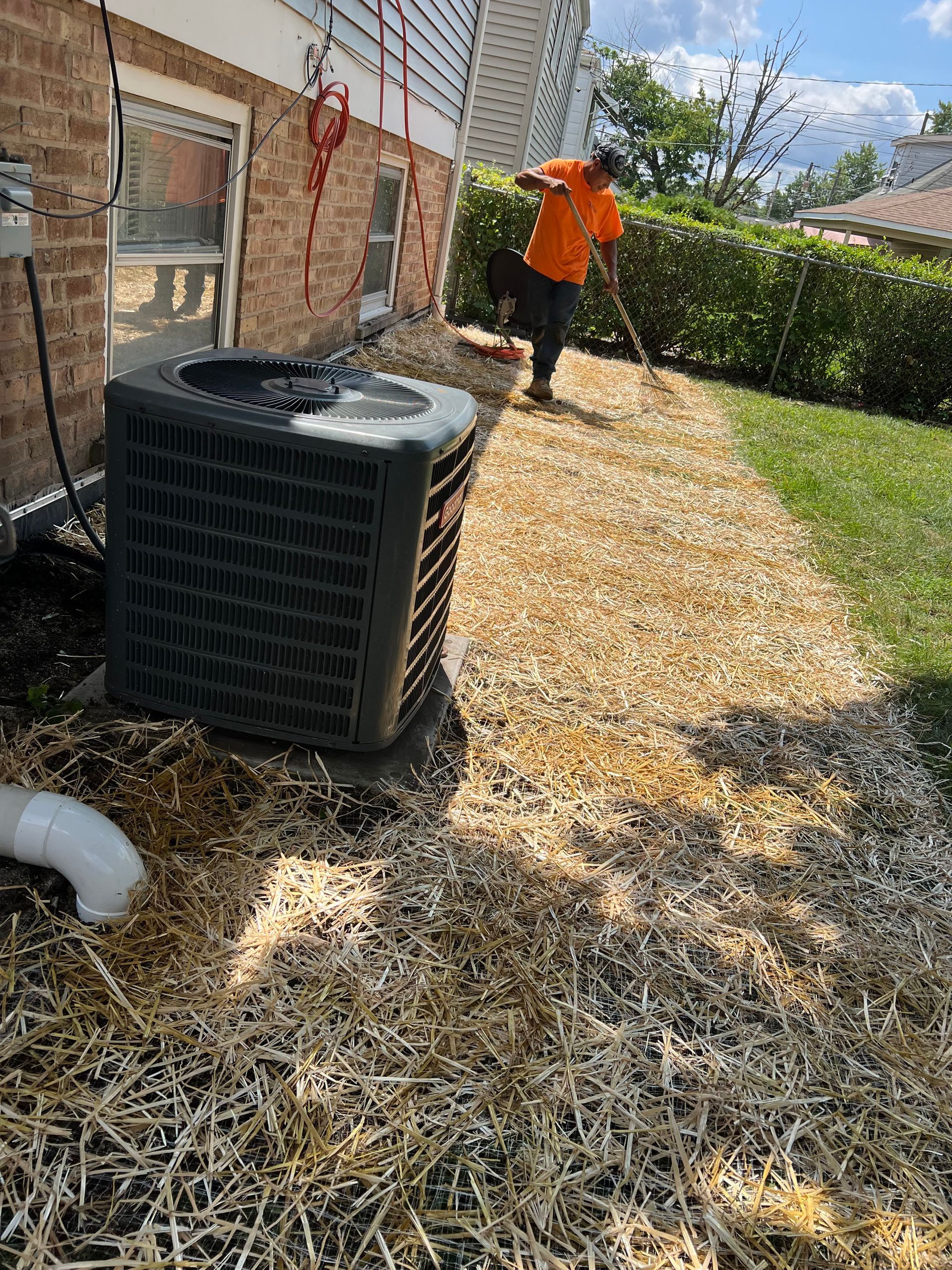 Person raking wood chips near a house and air conditioning unit. The setting is outdoors, with a brick wall and green grass.