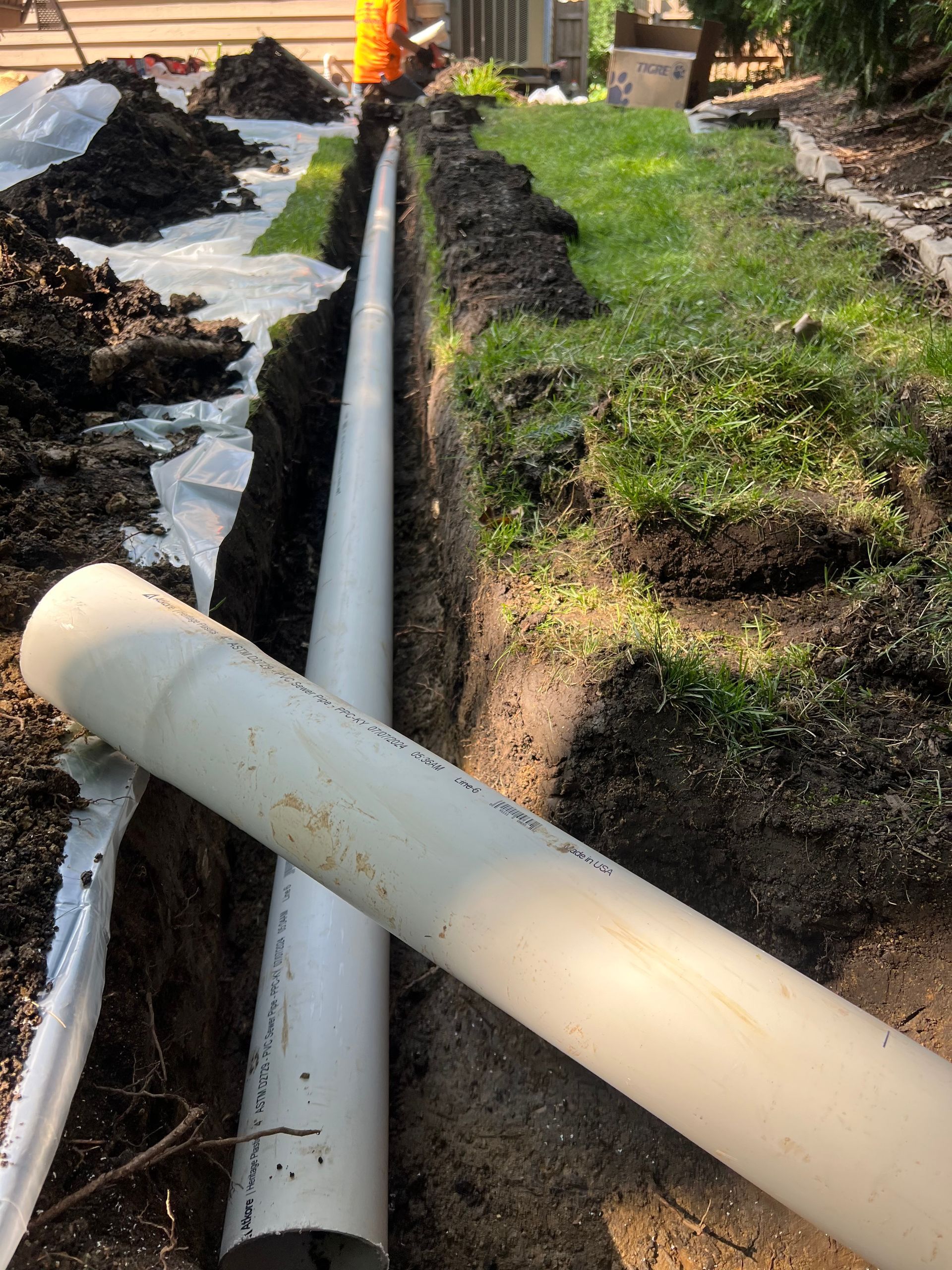 A trench holds a long, white pipe, with a second pipe lying beside it.  Dark soil and grass surround the trench.