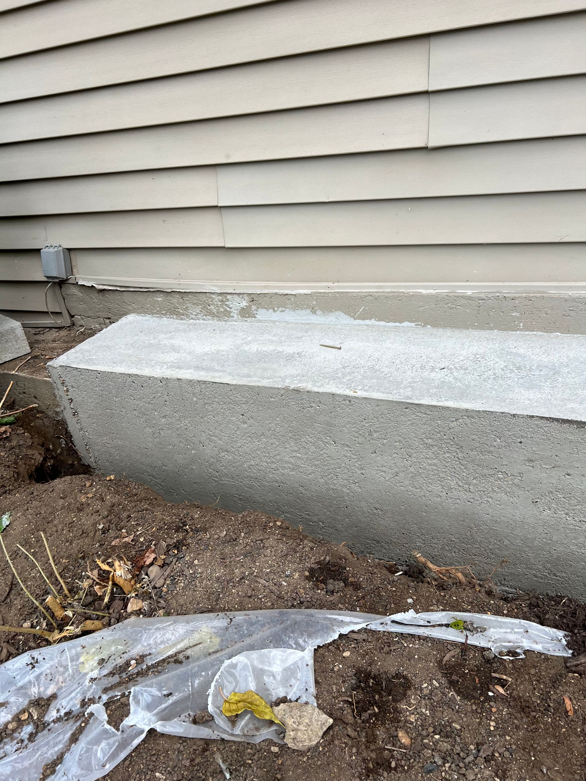 Concrete block foundation next to a building's siding, with soil and plastic sheeting in front.