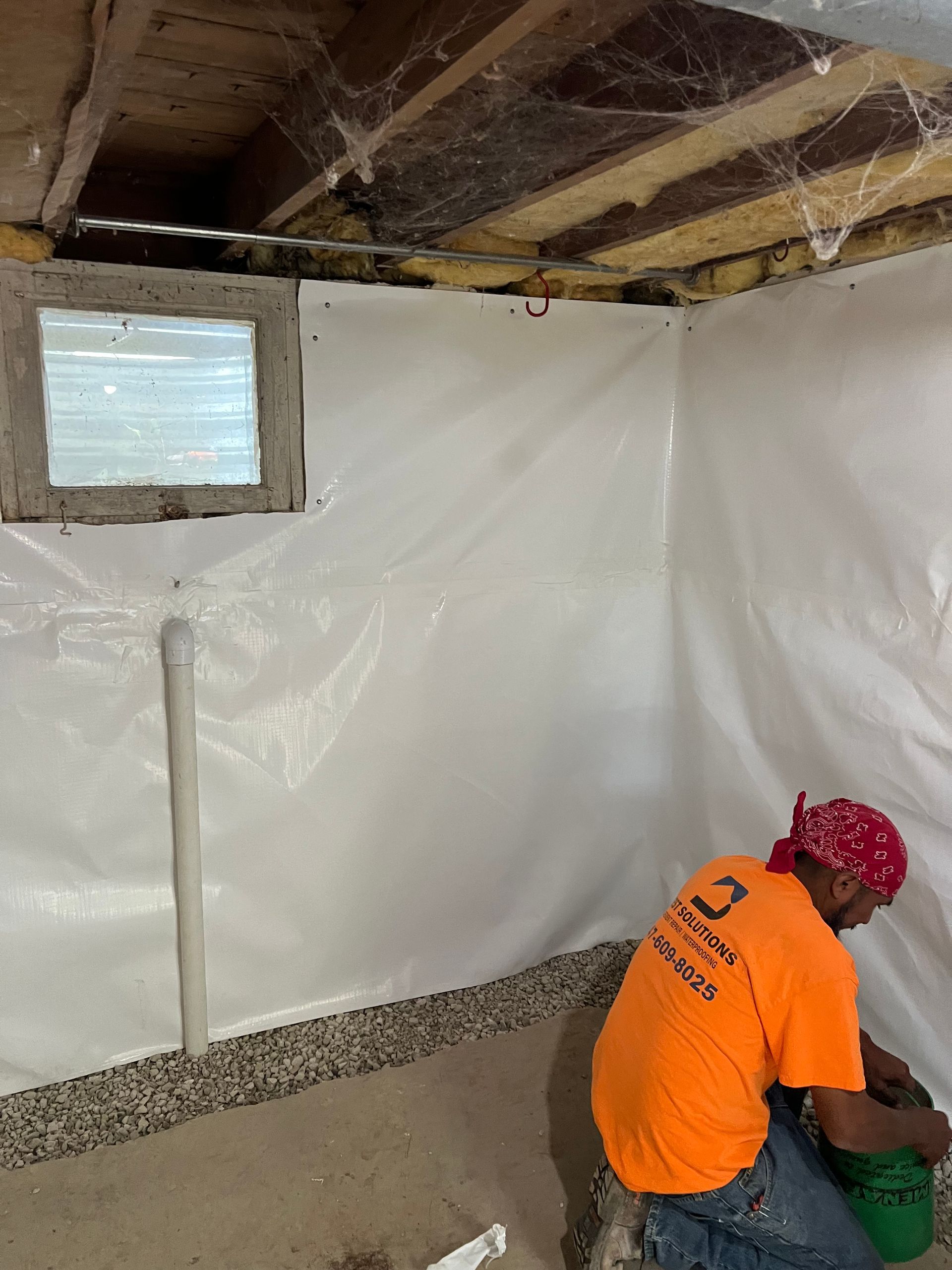 Man in orange shirt and red bandana working in a basement. White plastic covers the walls, cobwebs on the ceiling.