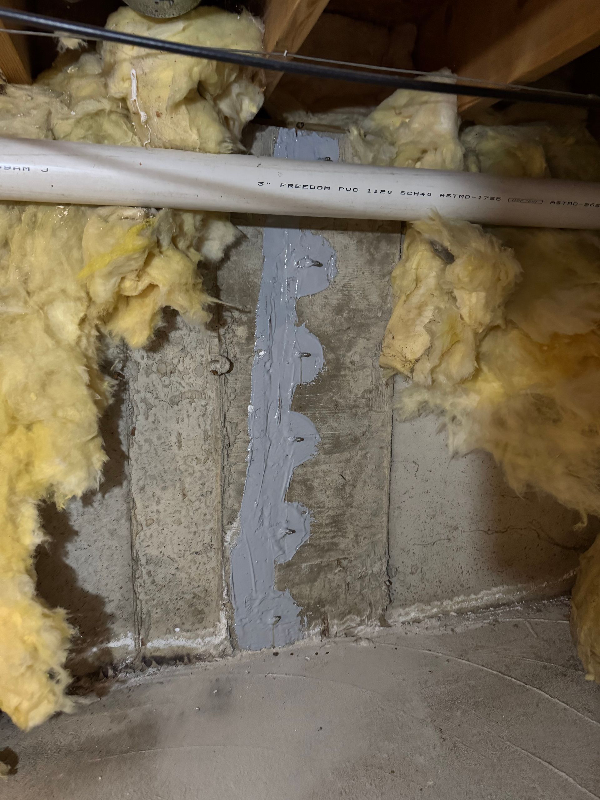 Concrete wall with a grey sealant applied vertically, surrounded by yellow insulation, in a crawl space setting.
