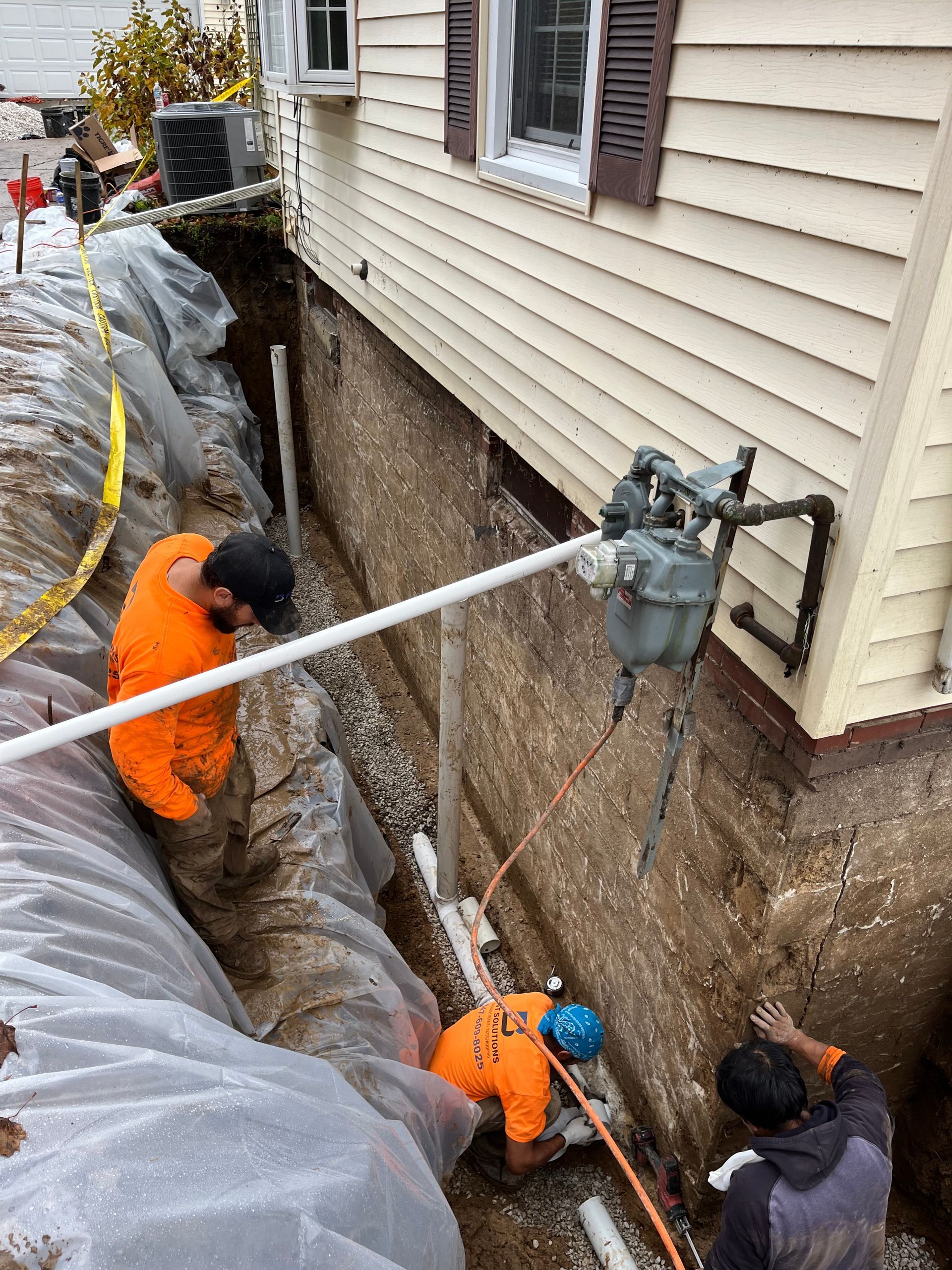 Three construction workers in orange coveralls are working in a deep trench next to a house. The ground is exposed, and plastic sheeting covers the trench.