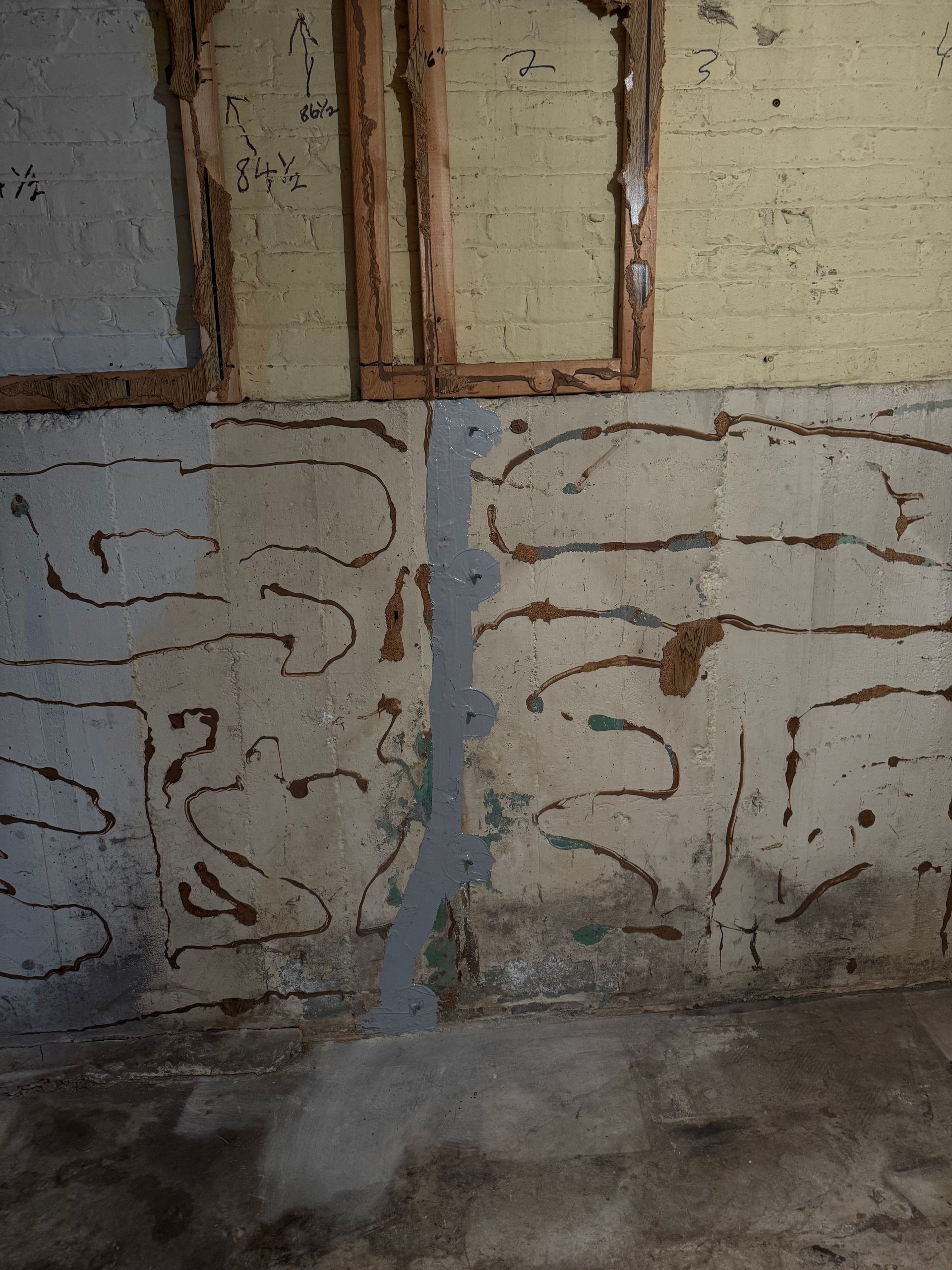 Concrete basement wall with a vertical gray patch and painted, splotchy, patterned sections. Above, are exposed wooden frames.