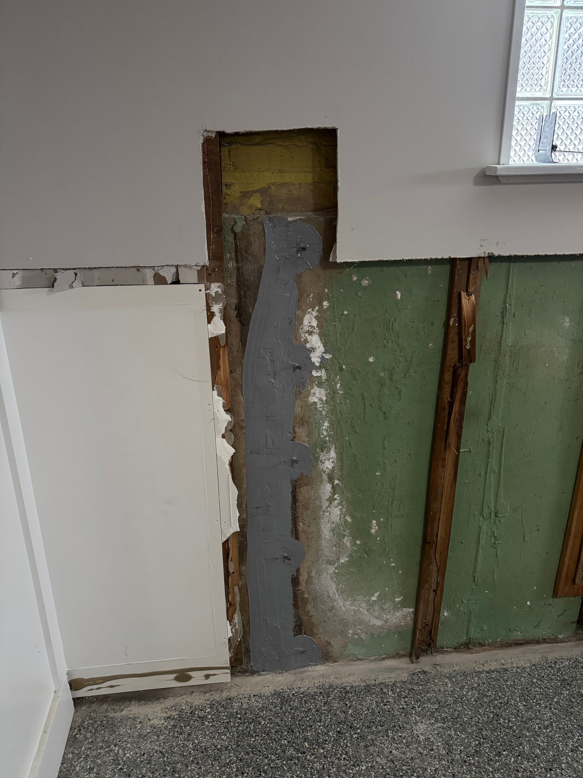 Wall with sections of damaged drywall, exposing wooden studs and a painted support. The walls are painted white, green, and grey.